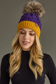 Woman wearing a purple and yellow knit beanie with a fur pom-pom on a gray background