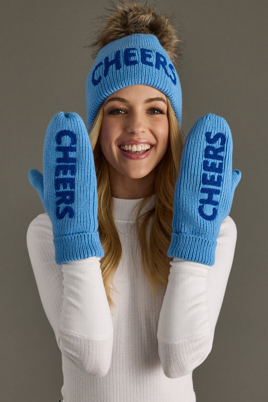 Woman wearing a blue 'CHEERS' beanie and mittens against a gray background