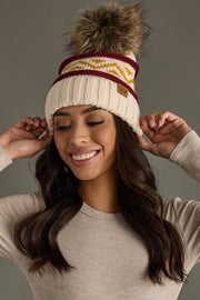 Woman wearing a cream beanie with a pom-pom and a logo, smiling against a gray background