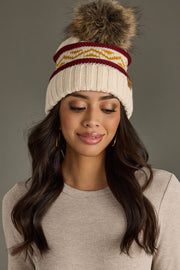 Woman wearing a cream knit beanie with colorful stripes and a fur pom-pom on a gray background