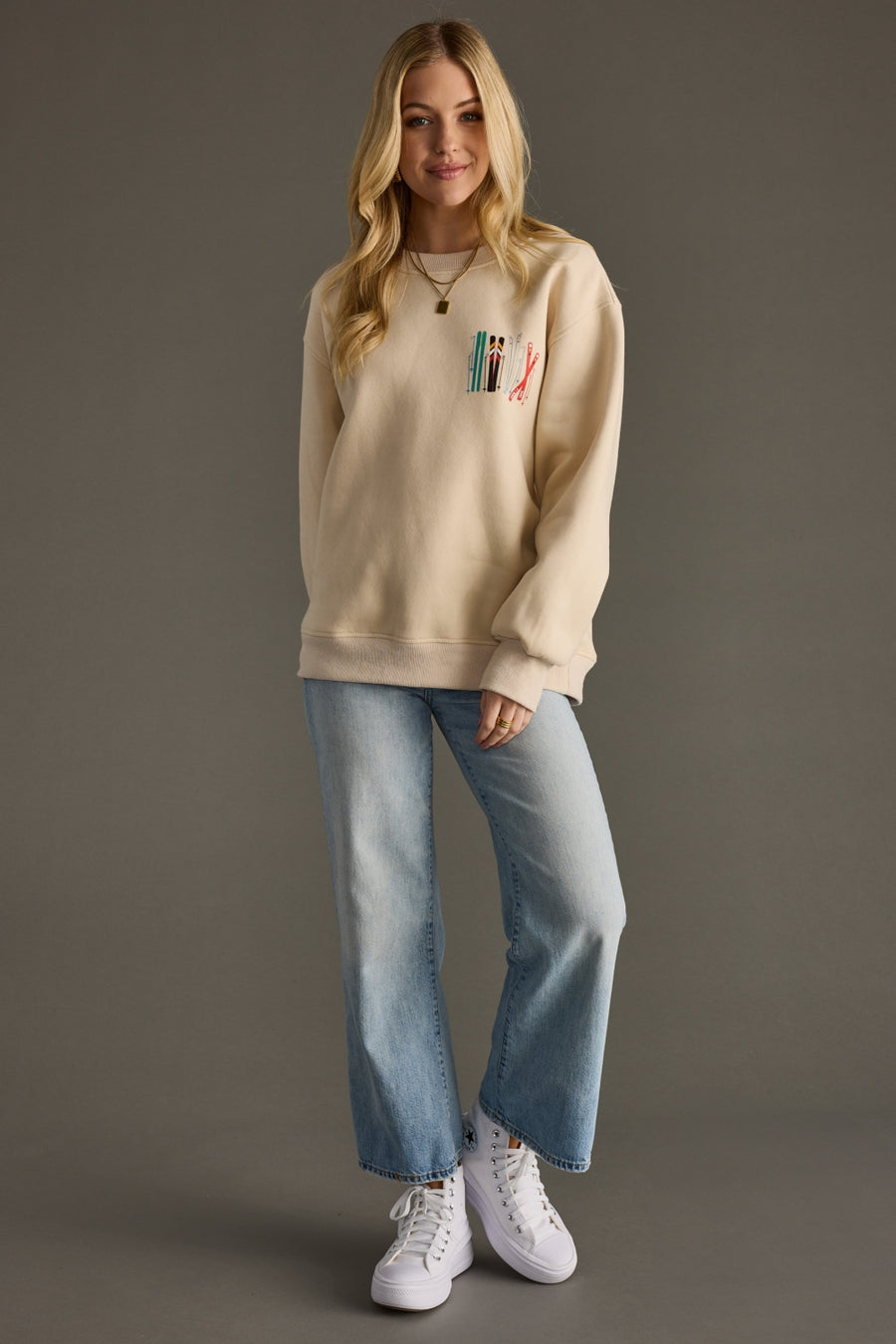 Woman wearing a beige sweatshirt with a graphic design, light blue jeans, and white sneakers on a gray background