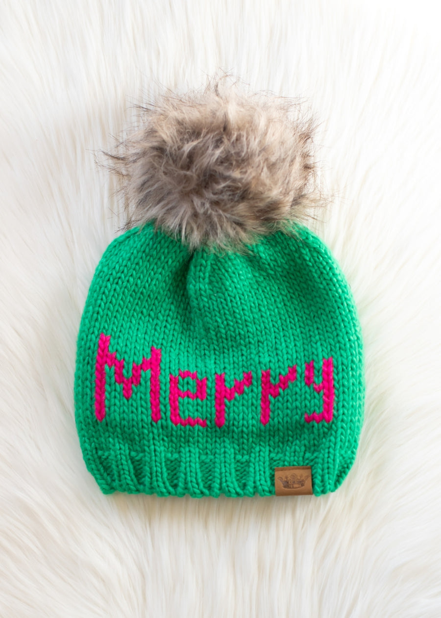 Green knit beanie with pink 'Merry' text and fur pom-pom on a light background