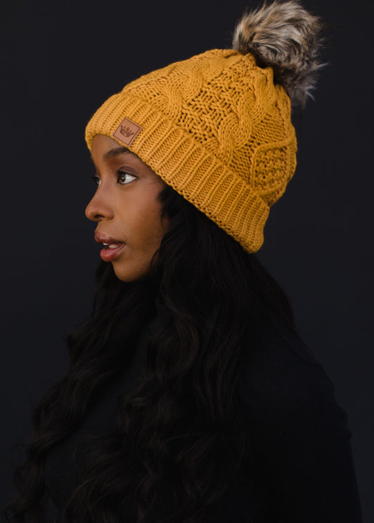 Woman wearing a mustard yellow knit beanie with a fur pom-pom on a black background