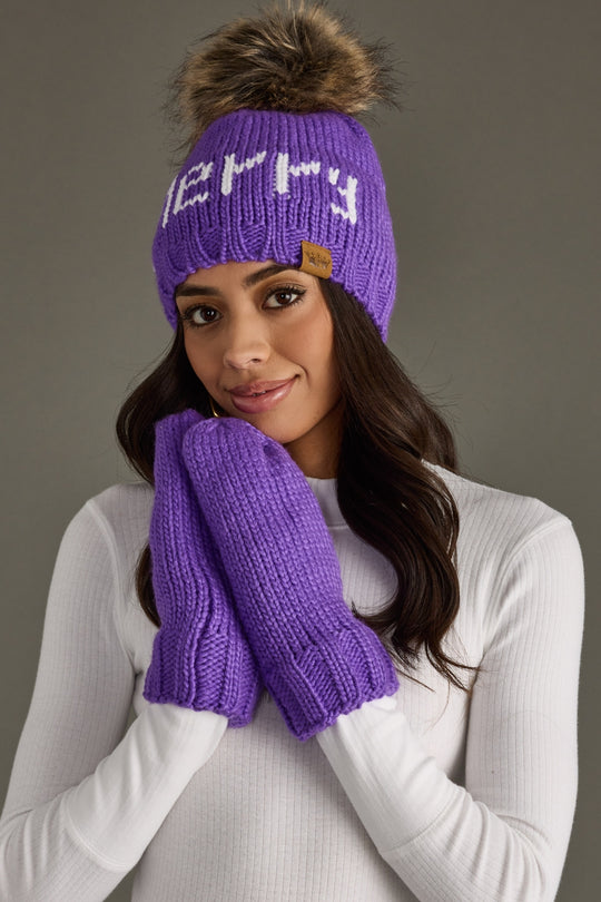 Woman wearing a purple knit beanie and mittens with a fur pom-pom on a gray background