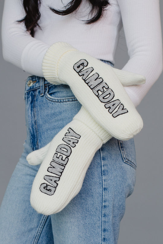 White 'GAMEDAY' gloves held by a person wearing a white sweater and blue jeans on a gray background