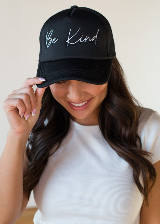 A woman with long brown hair wears a white short-sleeve top and smiles as she adjusts the Be Kind Trucker Hat by panacheaccessories—featuring "Be Kind" in white script and an adjustable snapback for comfort.