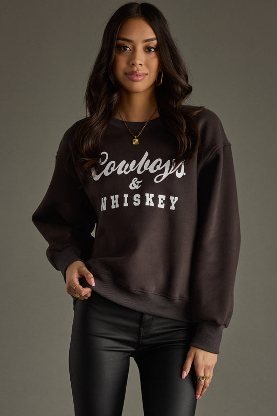 A woman with long dark hair poses indoors against a brick wall, wearing a charcoal Cowboys & Whiskey Sweatshirt by Panache Apparel Collection, paired with black shorts and boots, her hand resting confidently on a table.