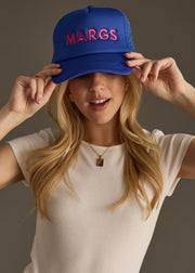 A woman in a fitted white top wears a bright blue baseball cap with bold pink text that reads “MARG.” She’s looking down with a soft smile, arms loosely crossed.