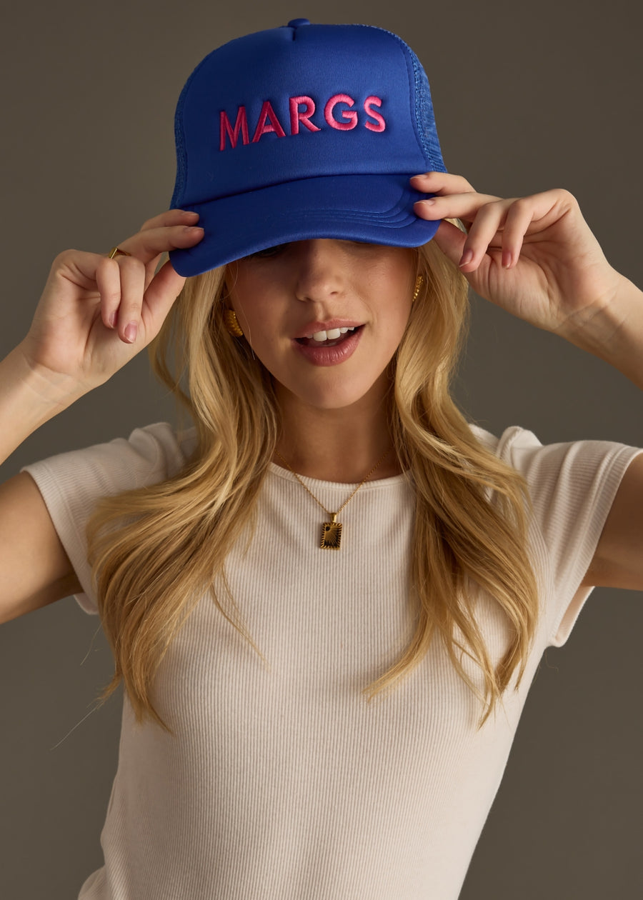 A woman in a fitted white top wears a bright blue baseball cap with bold pink text that reads “MARG.” She’s looking down with a soft smile, arms loosely crossed.