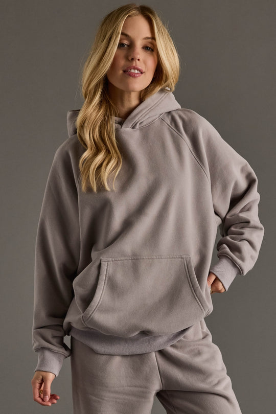 A woman with long blonde hair wears a Gray Panache Hooded Sweatshirt from the Panache Apparel Collection and matching sweatpants, standing against a plain gray background with one hand in her pocket.