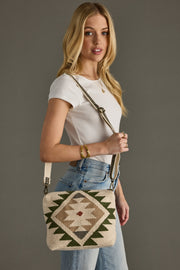 A woven crossbody bag features a striking geometric southwestern pattern with deep green triangular motifs and burgundy diamond accents on a natural beige textured base. The bag includes an adjustable crossbody strap with antique brass hardware, showcasing artisan craftsmanship with a bohemian-chic aesthetic against a rich teal background.