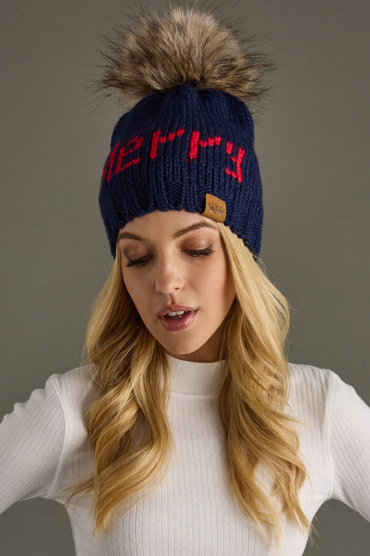 Woman wearing a blue knit beanie with a pom-pom and 'Merry' text, against a gray background