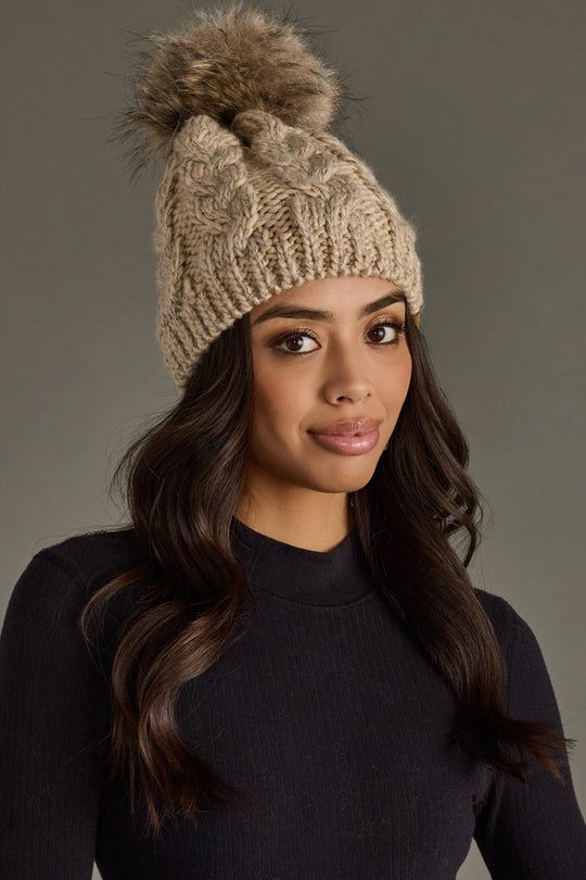 Woman wearing a beige knitted beanie with a fur pom-pom against a gray background