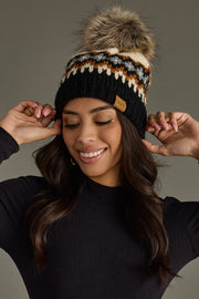 Woman wearing a black beanie with a fur pom-pom, smiling against a gray background