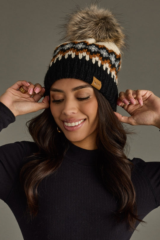 Woman wearing a black beanie with a fur pom-pom, smiling against a gray background