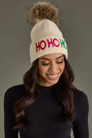 Woman wearing a cream beanie with 'HO HO HO' in colorful letters and a fur pom-pom on a gray background.