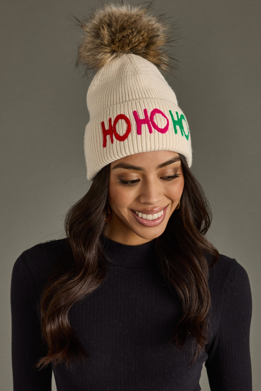 Woman wearing a cream beanie with 'HO HO HO' in colorful letters and a fur pom-pom on a gray background.