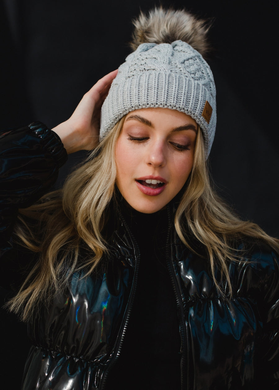 A woman with long blonde hair smiles while looking slightly to the side, wearing a light gray knit beanie with a faux-fur pom-pom and a small leather patch on the front. She’s dressed in a dark, glossy winter jacket, posed against a plain dark background that highlights her warm expression and cozy cold-weather style.