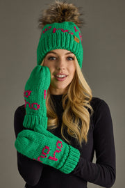 A smiling woman wears the Panache Apparel Collection Ho Ho Ho Pom Hat - Green & Pink, paired with a black long-sleeve top, posing against a plain grey background.