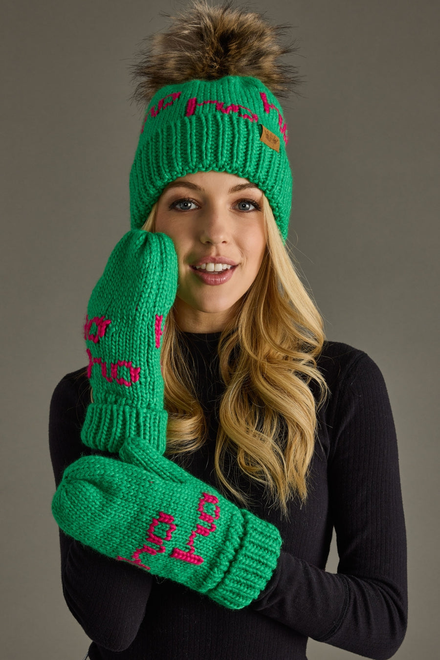 A smiling woman wears the Panache Apparel Collection Ho Ho Ho Pom Hat - Green & Pink, paired with a black long-sleeve top, posing against a plain grey background.