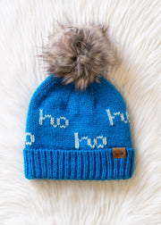 The Ho Ho Ho Pom Hat - Blue by Panache Apparel Collection features bright blue fleece lining, a large faux fur pom-pom, white “ho ho ho” text, and a brown leather tag, shown on a fluffy white surface.