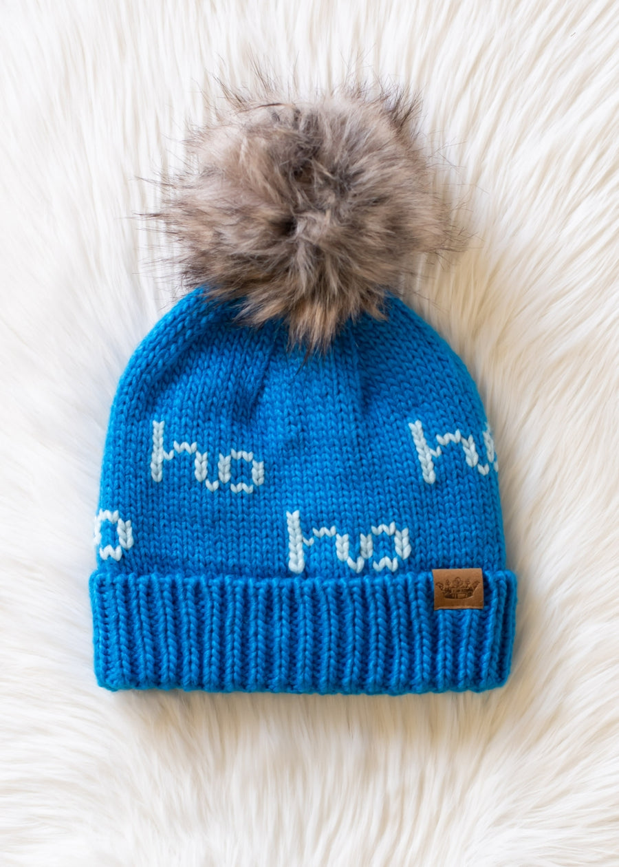 The Ho Ho Ho Pom Hat - Blue by Panache Apparel Collection features bright blue fleece lining, a large faux fur pom-pom, white “ho ho ho” text, and a brown leather tag, shown on a fluffy white surface.