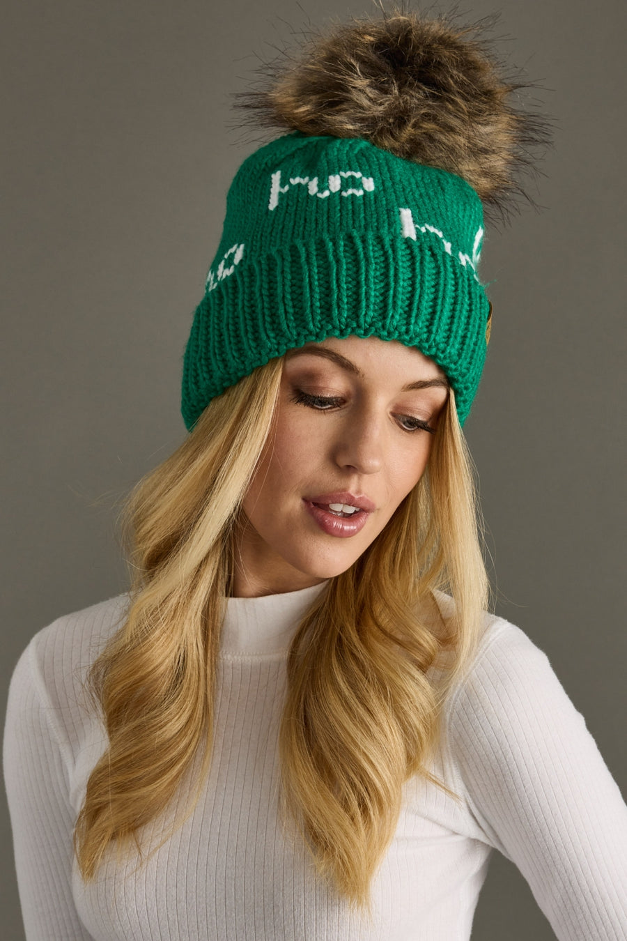 A woman with long blonde hair wears the Panache Apparel Collection's Ho Ho Ho Pom Hat in green and white, featuring a large faux fur pom and cozy fleece lining, paired with a white ribbed turtleneck as she looks down with a neutral expression.