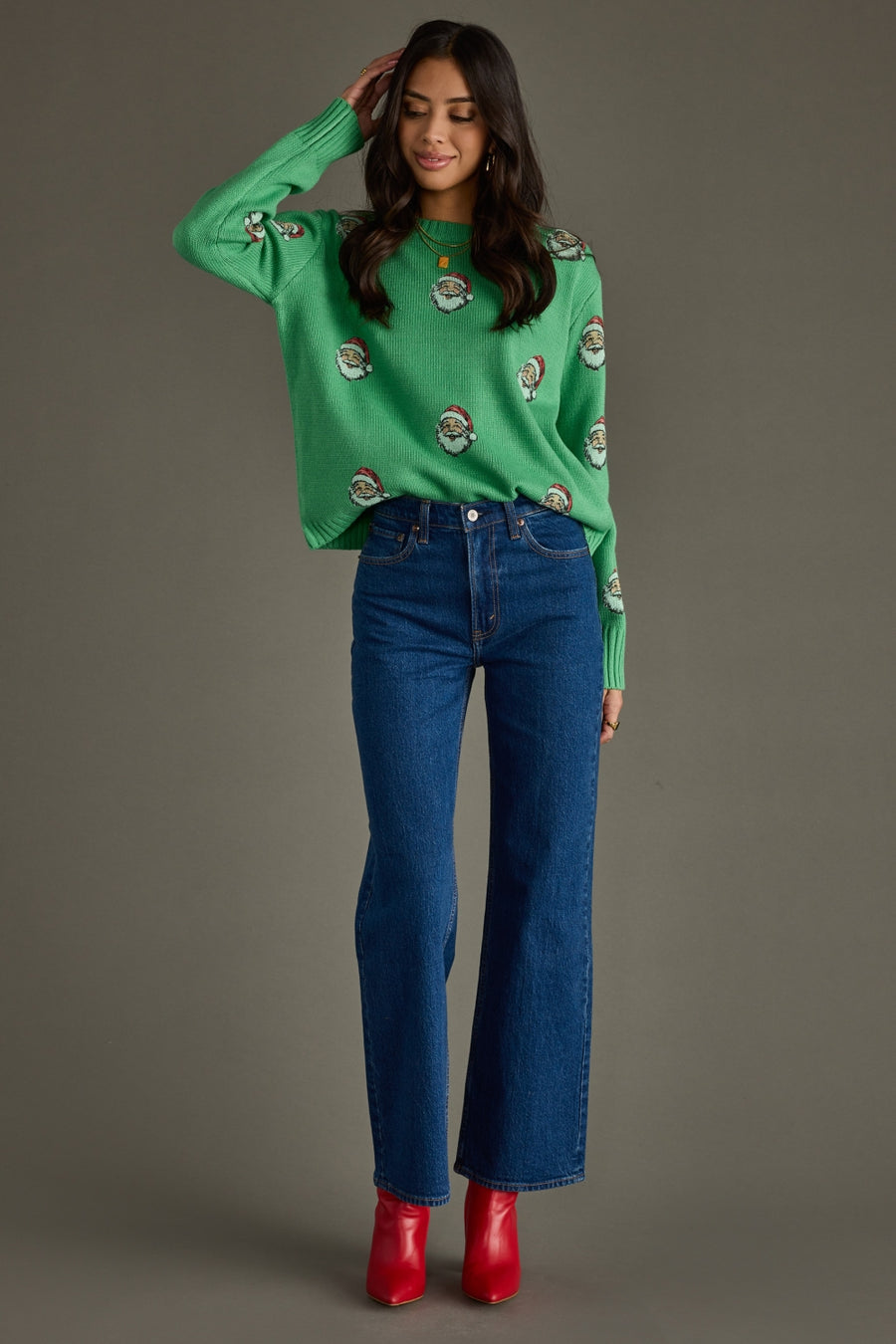 Woman wearing a green sweater with Santa pattern, blue jeans, and red shoes on a gray background