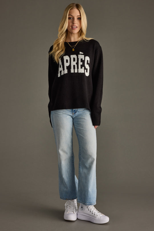 Woman wearing a black sweater with 'APRèS' text, light blue jeans, and white sneakers on a gray background