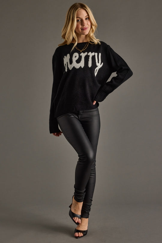 A woman stands against a gray backdrop, smiling with one hand on her hip and one foot slightly raised, wearing the Panache Apparel Collection Black Merry Sweater—a relaxed fit black knit featuring "merry" in white—paired with black faux leather leggings and black heels.