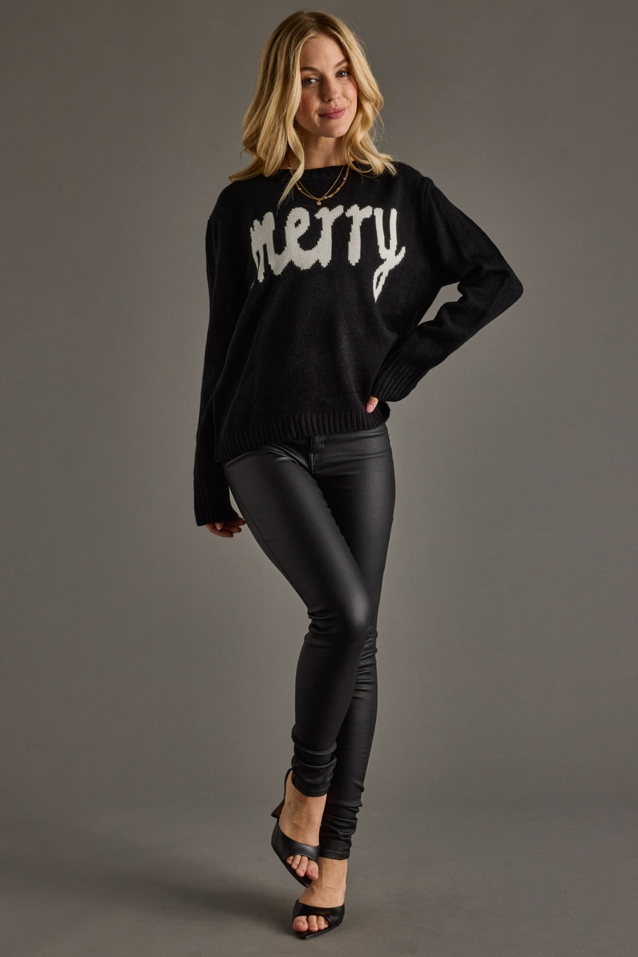 Woman wearing a black sweater with 'merry' text, black pants, and high heels on a gray background