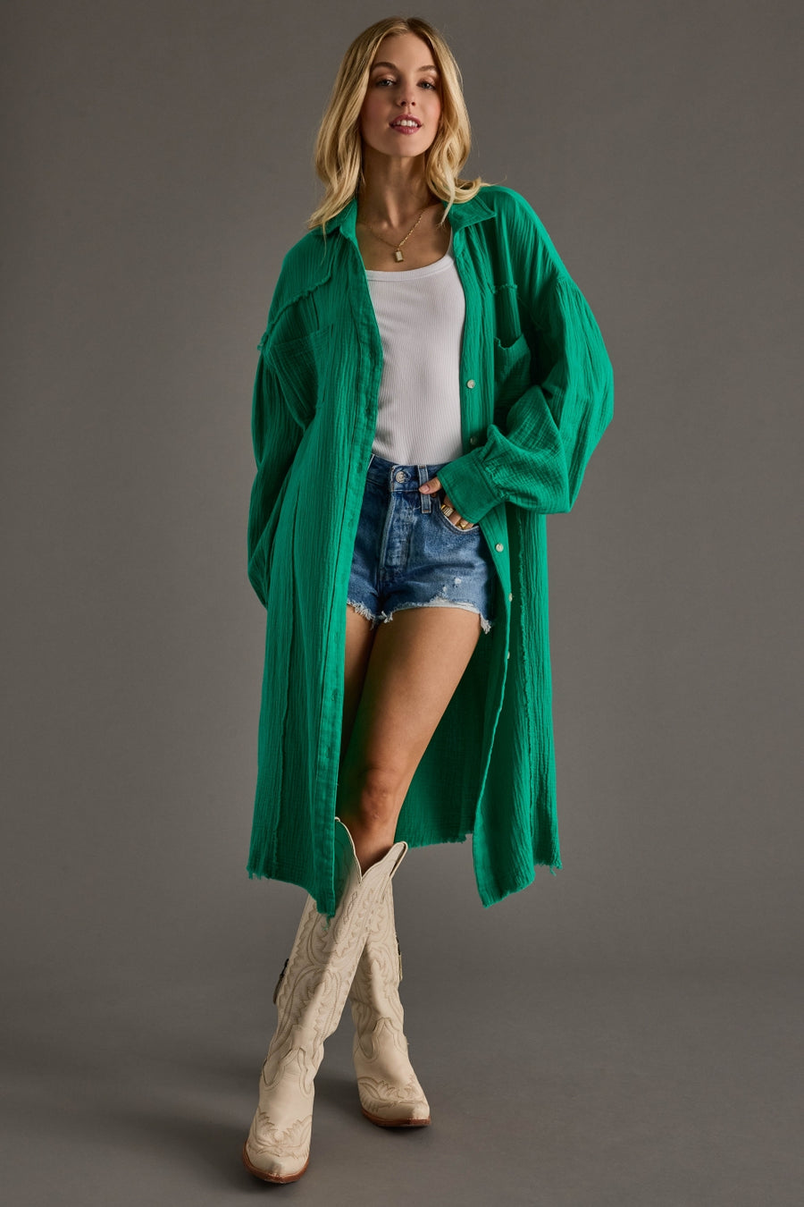 Woman wearing a green cardigan, white top, denim shorts, and beige boots on a gray background