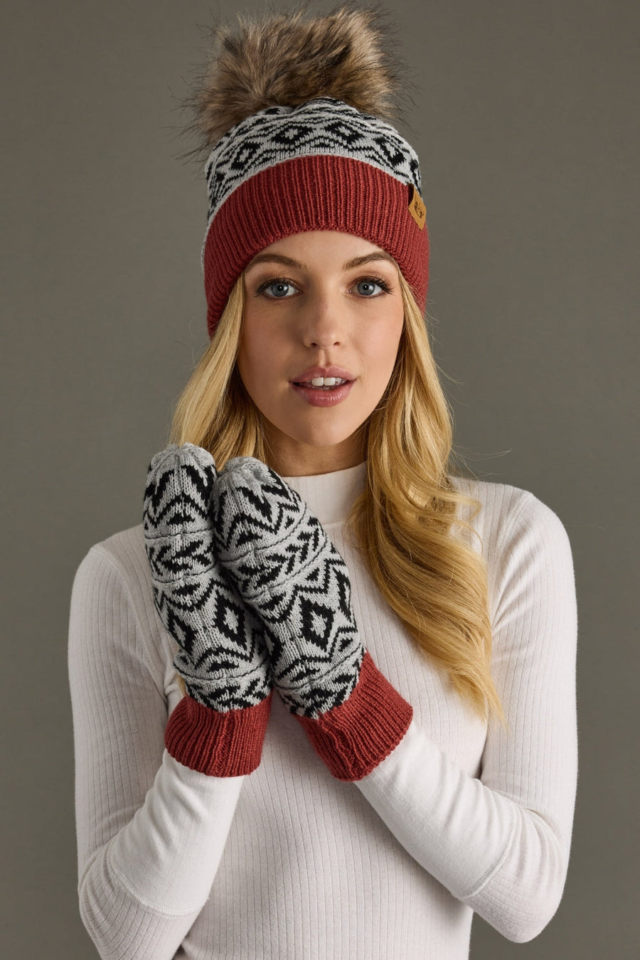 A knit beanie features a classic Fair Isle geometric pattern in cream and black with a solid rust-red ribbed cuff and small tan leather logo patch. Topped with a luxurious faux-fur pom-pom, this winter hat delivers Nordic-inspired alpine style with cozy warmth.