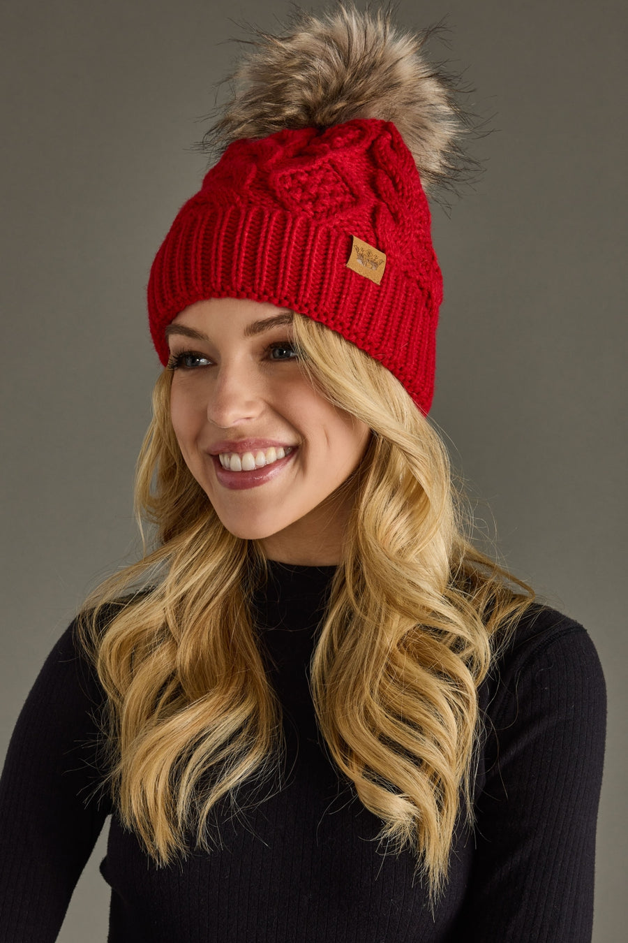 A smiling woman with long blonde hair models the Panache Apparel Collection Red Cable Knit Pom Hat, featuring a faux fur pom-pom, paired with a black sweater and set against a plain gray background.