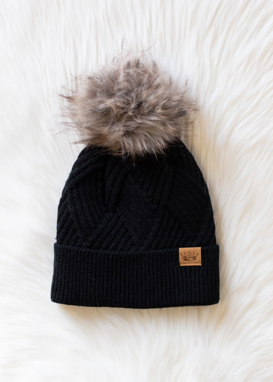 Black knit beanie with fur pom-pom on a white textured background