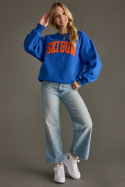 Woman wearing a blue sweatshirt with 'Ski bum' text, light blue jeans, and white sneakers on a gray background