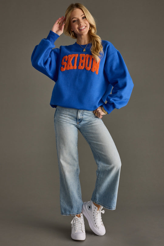 Woman wearing a blue sweatshirt with 'Ski bum' text, light blue jeans, and white sneakers on a gray background