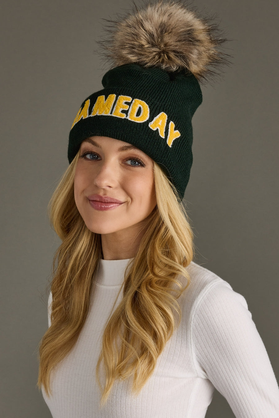Woman wearing a green beanie with 'GAME DAY' text and fur pom-pom on a gray background