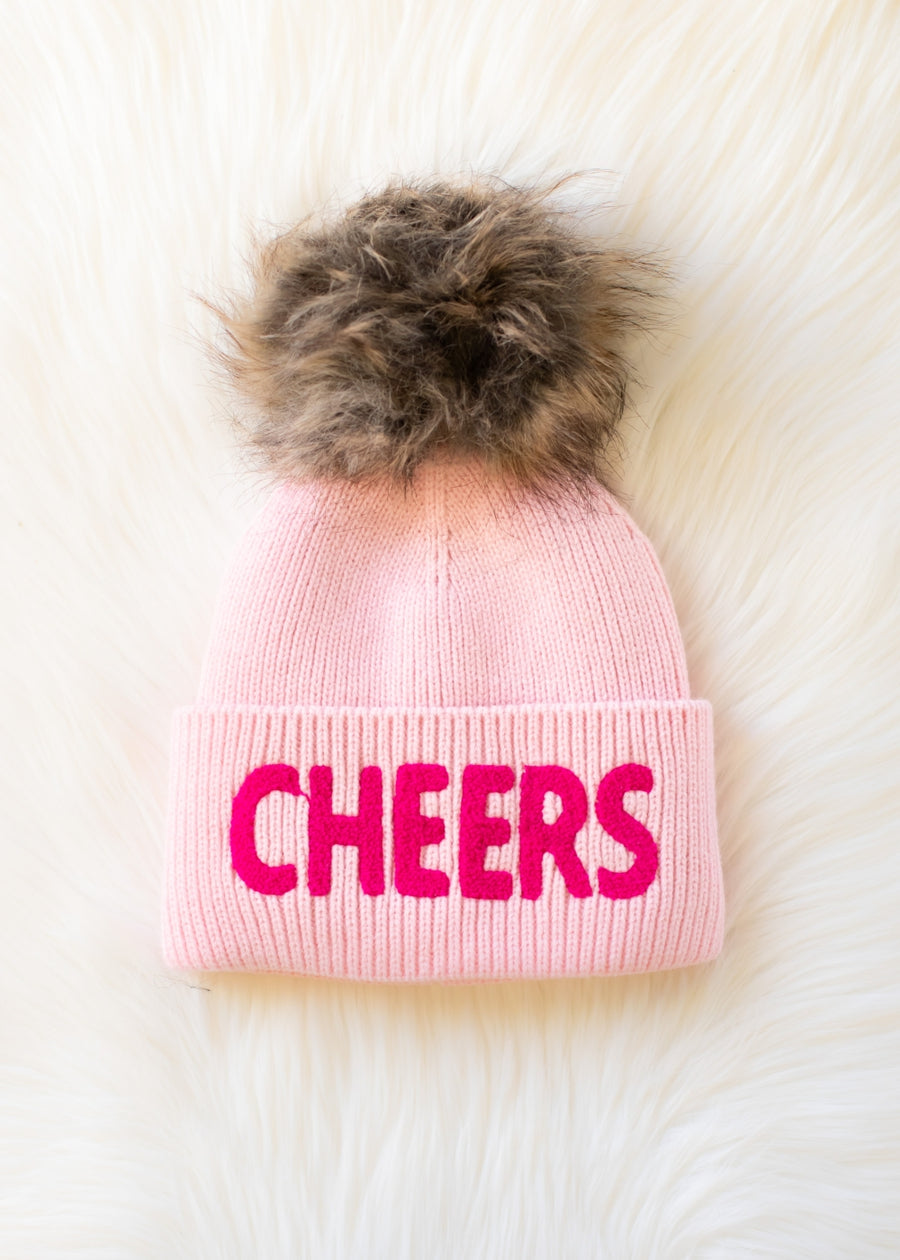 The Cheers Pom Hat - Pink by Panache Apparel Collection features light pink fleece-lined knit, a brown faux fur pom-pom, and bold bright pink "CHEERS" lettering on the front. The beanie is displayed on a fluffy white surface.