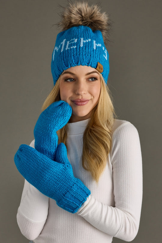 Woman wearing a blue knit hat with a pom-pom and matching gloves against a gray background