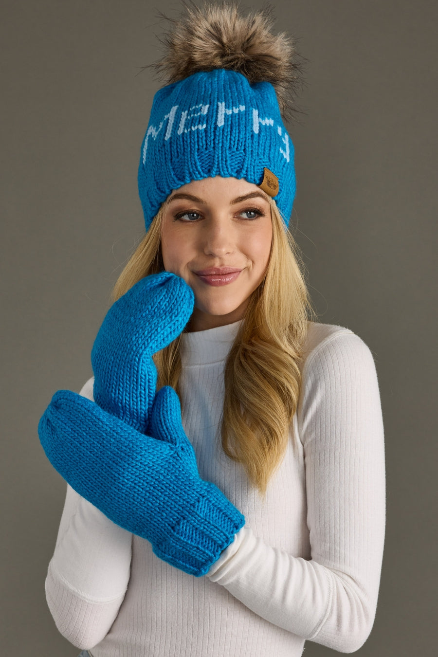 Woman wearing a blue knit hat with a pom-pom and matching gloves against a gray background