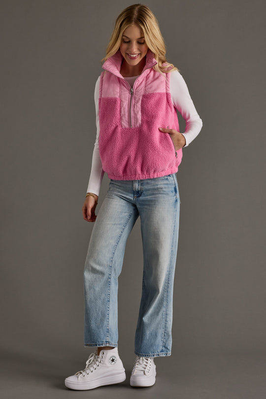 Woman wearing a pink fleece vest over a white long-sleeve shirt, blue jeans, and white sneakers on a gray background.