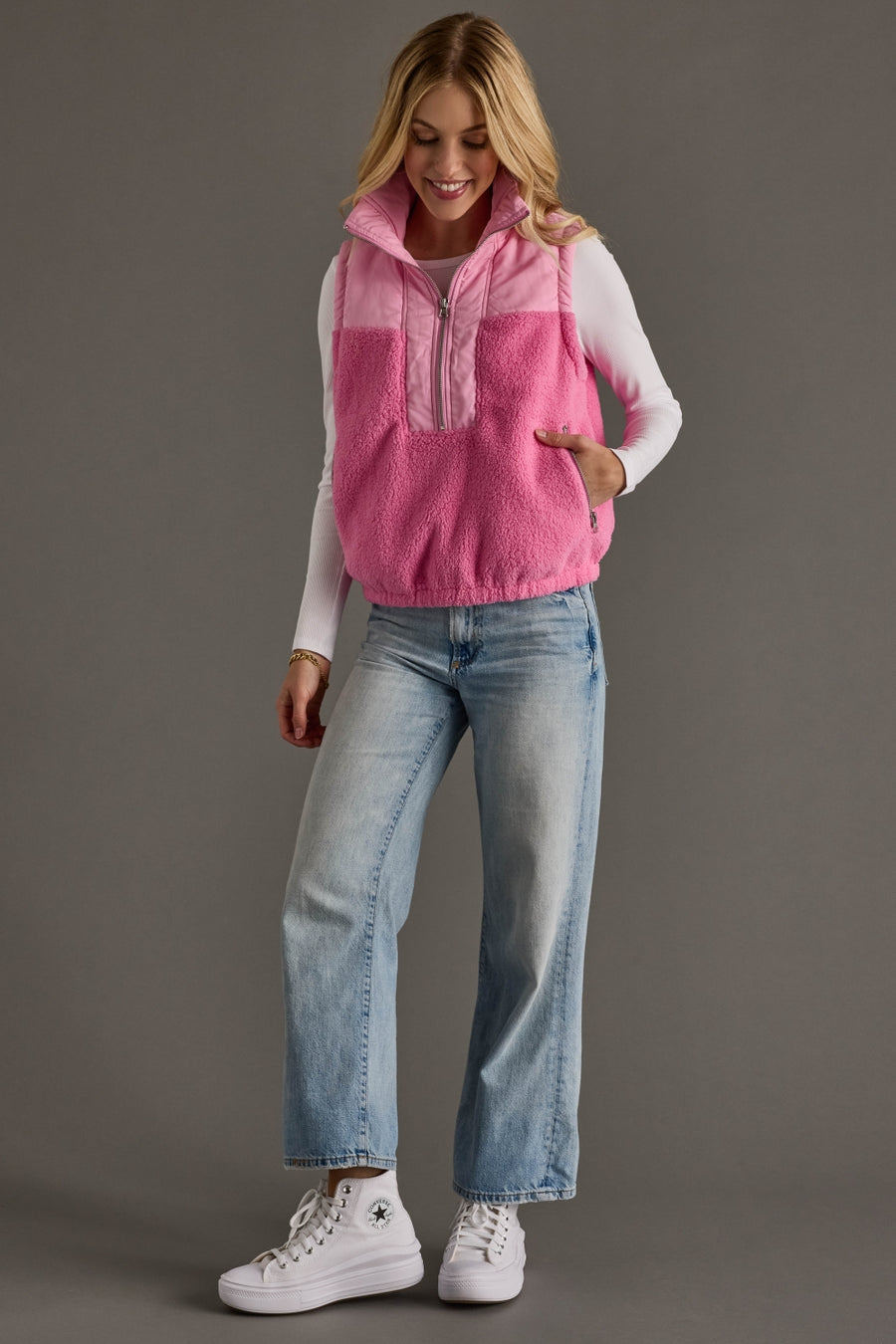 Woman wearing a pink fleece vest over a white long-sleeve shirt, blue jeans, and white sneakers on a gray background.