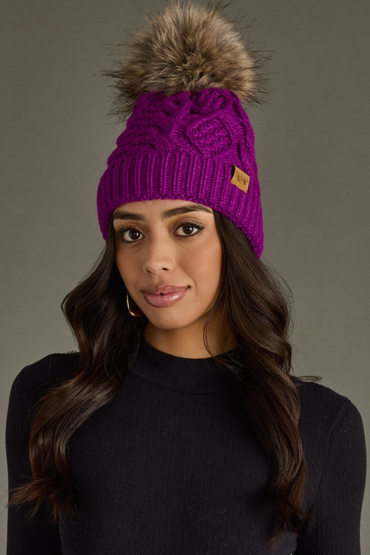 Woman wearing a purple knit beanie with a fur pom-pom against a gray background