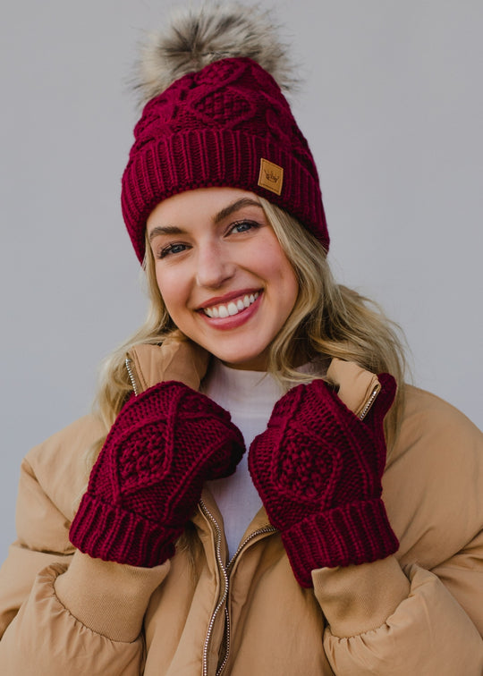 Woman wearing a red knit beanie and mittens with a beige coat against a gray background