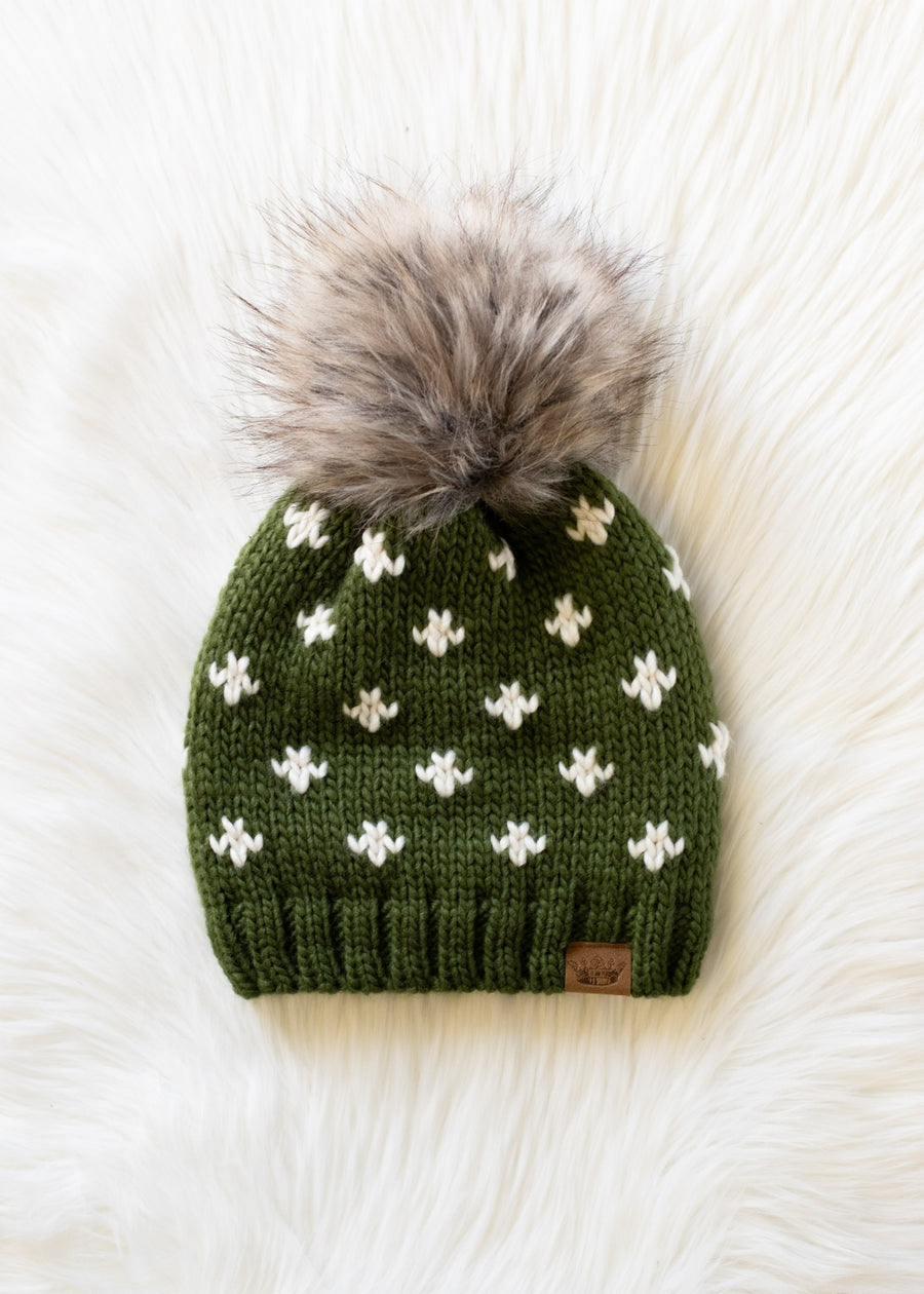 Green knit beanie with white patterns and a gray pom-pom on a white fur background
