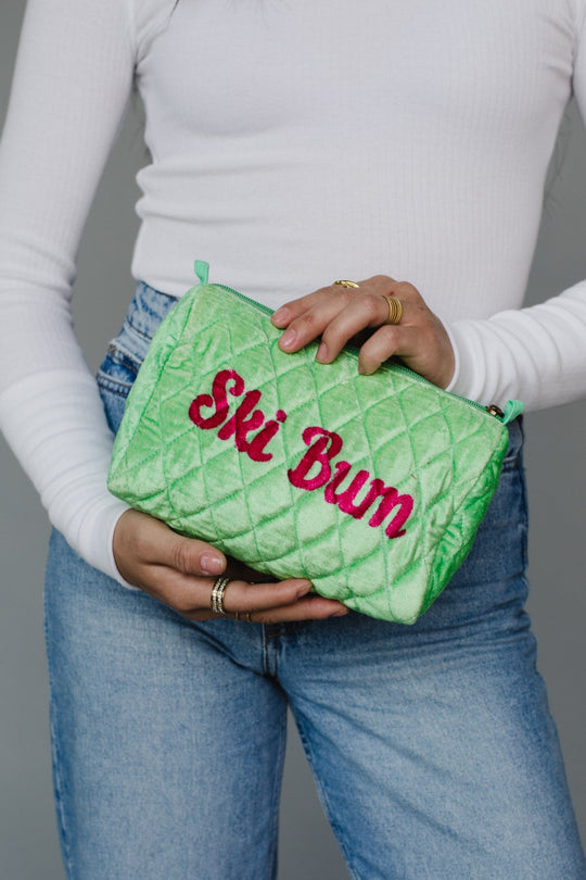 Person holding a green quilted pouch with 'Ski Bum' text against a gray background
