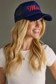 A smiling woman with long blonde hair wears the Panache Apparel Collection Merry Trucker Hat in navy, styled with a white ribbed tee and gold necklace. She looks sideways, highlighting the hat's adjustable snapback against a gray background.