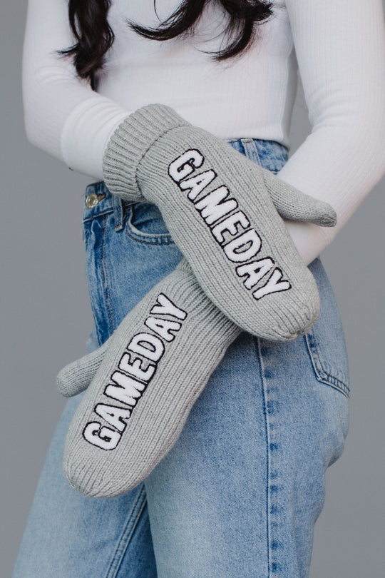Gray 'GAMEDAY' mittens worn by a person with a white sweater and blue jeans on a gray background