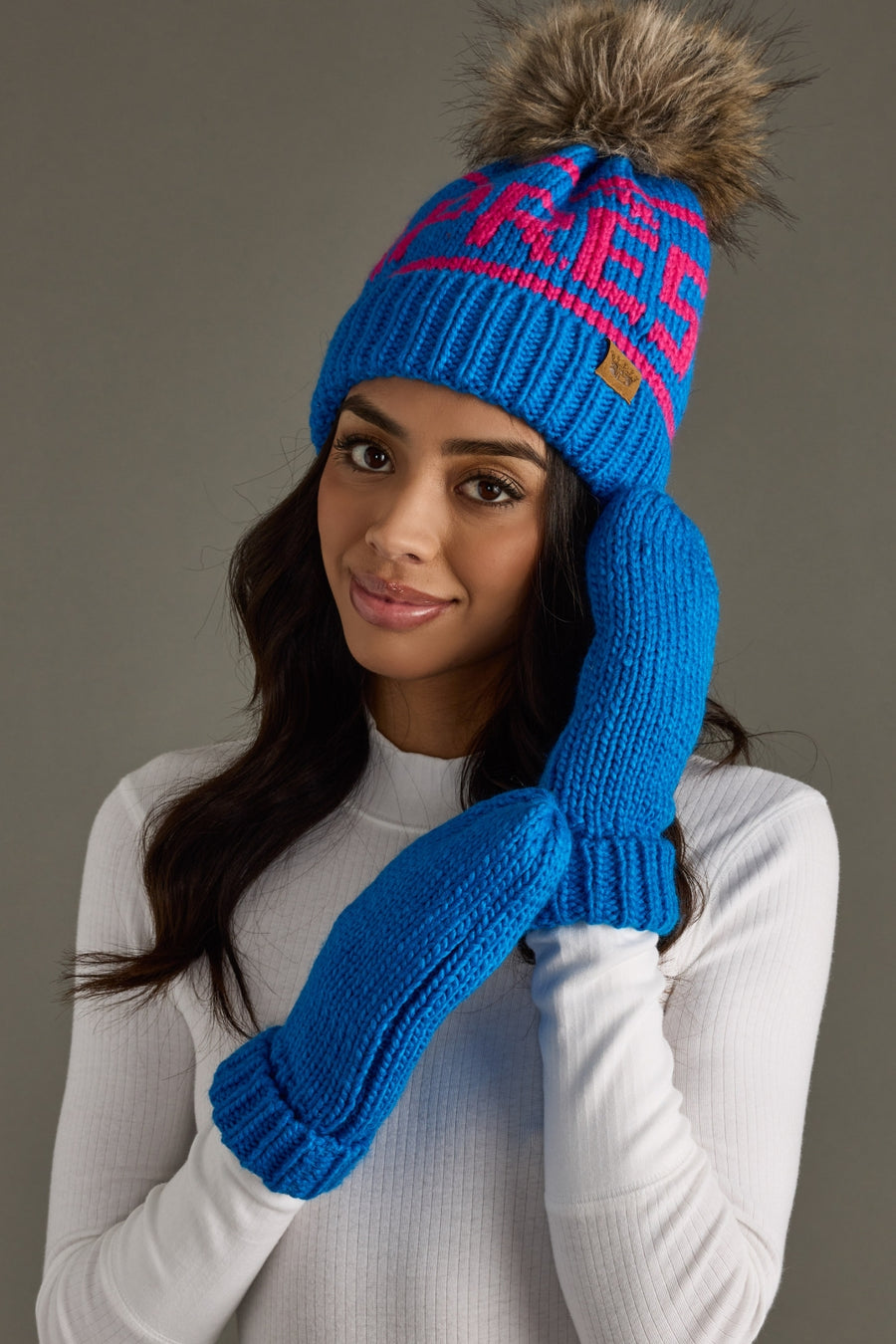 Woman wearing a blue knit hat and mittens with a visible brand logo on a gray background